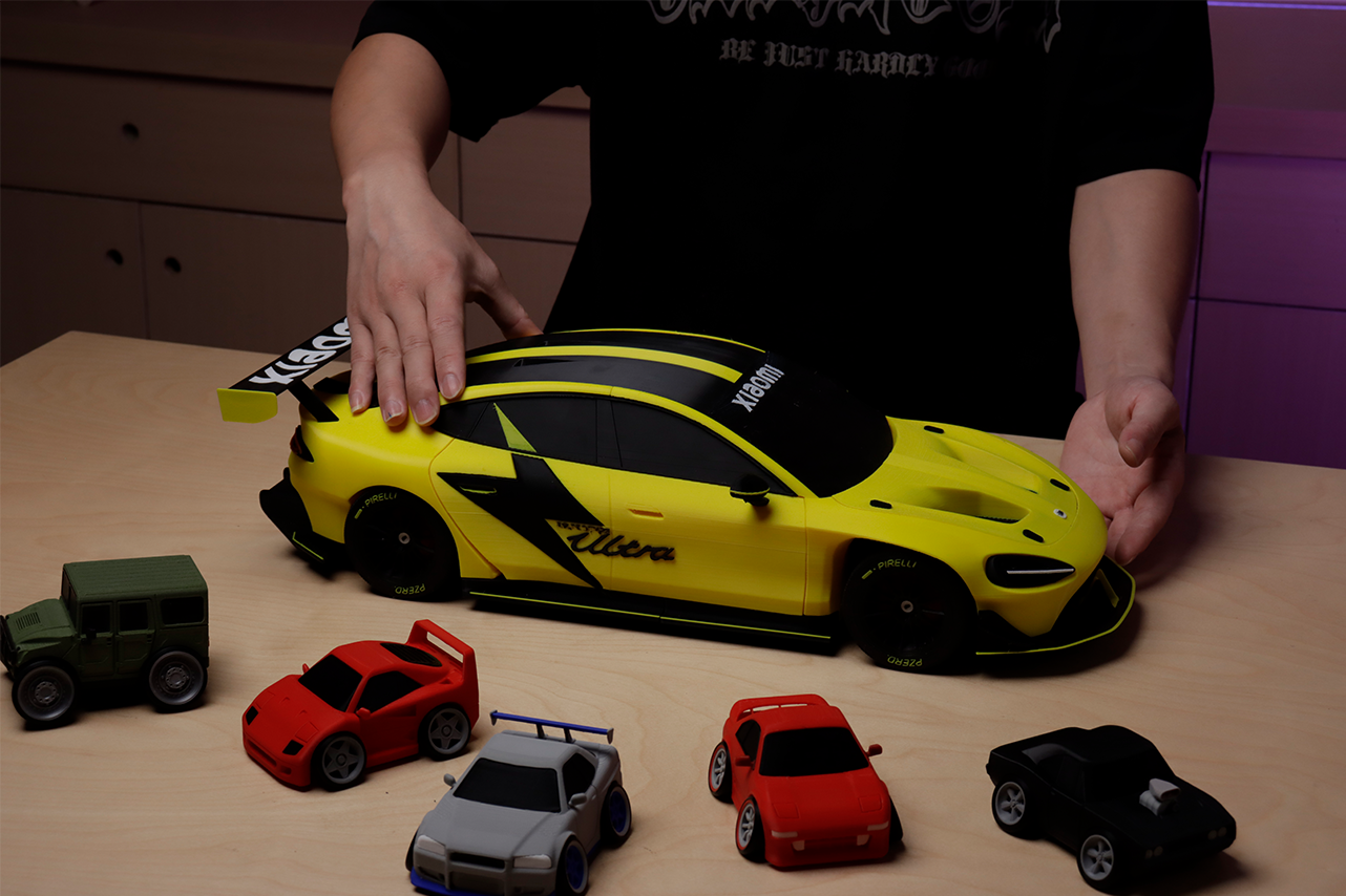 Hands assembling a large yellow and black toy car, surrounded by smaller colorful cars, showcasing a multi - color 3D printed car project that can be part of creative or functional 3D printing ideas.