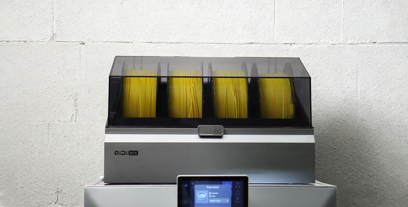 A silver QIDI BOX filament dryer holding four spools of yellow filament, with its control panel in the foreground